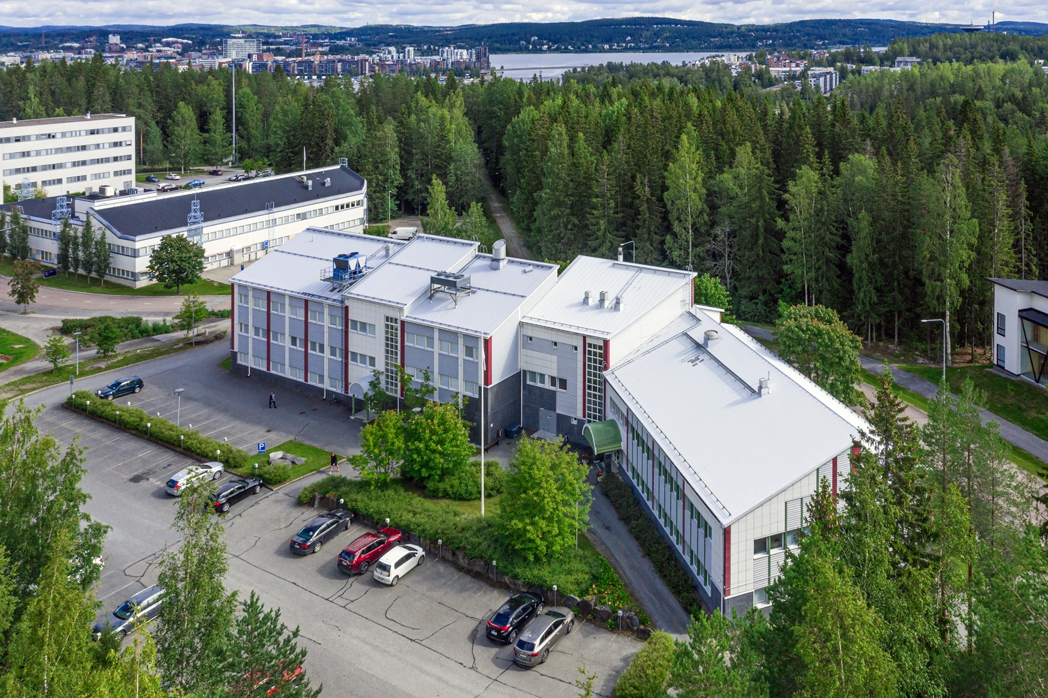 Ilmakuvassa 3-kerroksinen viistokattoinen toimitilarakennus Ylistönmäentie 31. Taustalla näkyy metsää, Jyväsjärvi ja Jyväskylän keskusta. Talon edessä pysäköintialue, jolla autoja.